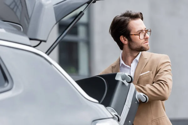 Homem distraído em óculos e terno colocando bagagem no porta-malas do carro embaçado — Fotografia de Stock