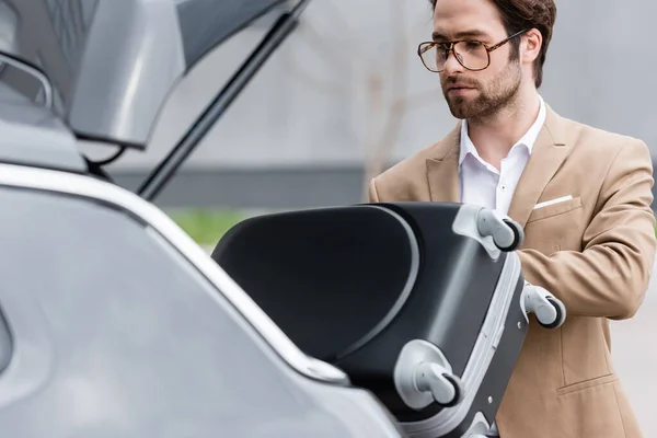 Bearded man in glasses and suit putting baggage in blurred car trunk — Stock Photo