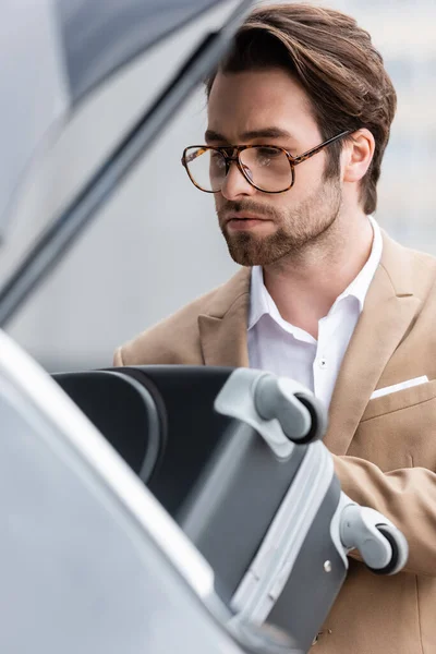 Bearded man in glasses and suit putting suitcase in blurred car trunk — Stock Photo