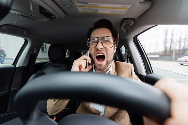 Сердитий чоловік говорить на смартфоні під час водіння автомобіля на розмитому передньому плані — стокове фото