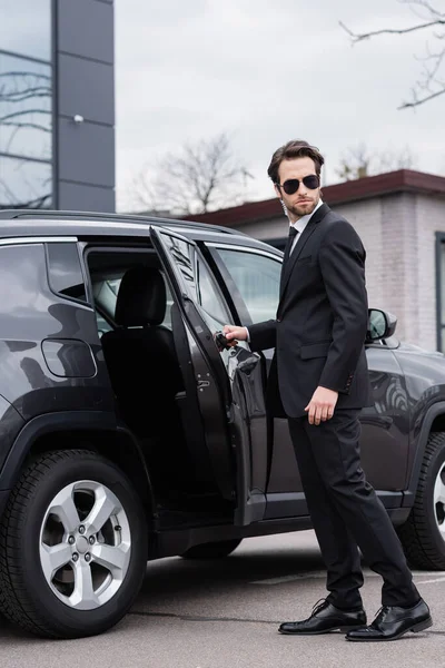 Bearded bodyguard in suit and sunglasses with security earpiece opening car door — Stock Photo