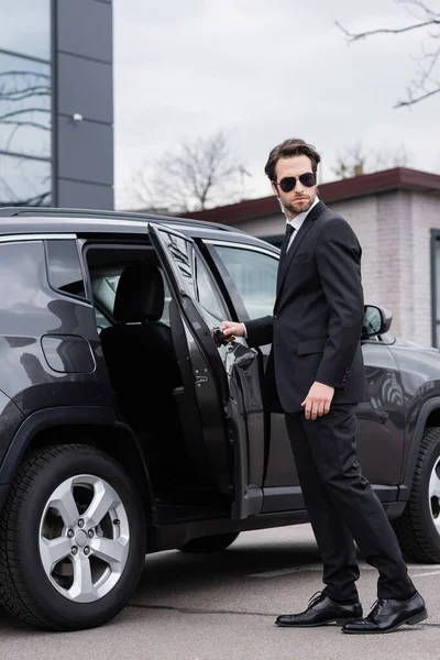 Garde du corps barbu en costume et lunettes de soleil avec écouteur de sécurité ouvrant la porte de la voiture — Photo de stock