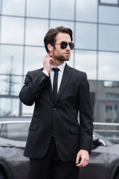 Guardaespaldas barbudo en traje y gafas de sol con auricular de seguridad cerca de edificio moderno sobre fondo borroso - foto de stock