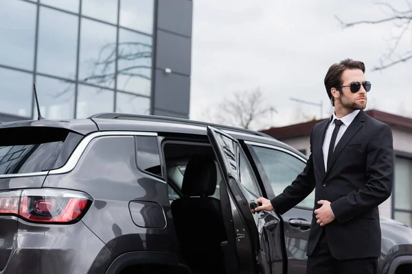 Bearded safeguard in suit and sunglasses with security earpiece opening car door — Stock Photo