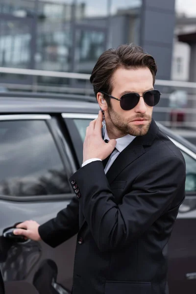 Bearded safeguard in suit and sunglasses adjusting security earpiece while opening car door — Stock Photo