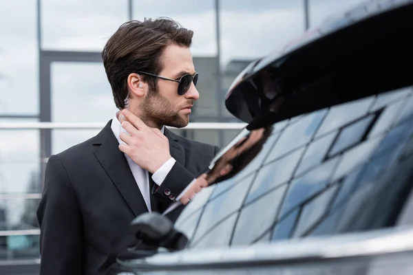 Garde du corps barbu en costume et lunettes de soleil ajustant écouteur de sécurité près de la voiture floue — Photo de stock