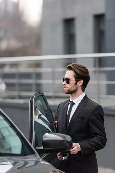 Hombre barbudo en traje y gafas de sol de pie cerca de coche moderno con puerta abierta - foto de stock