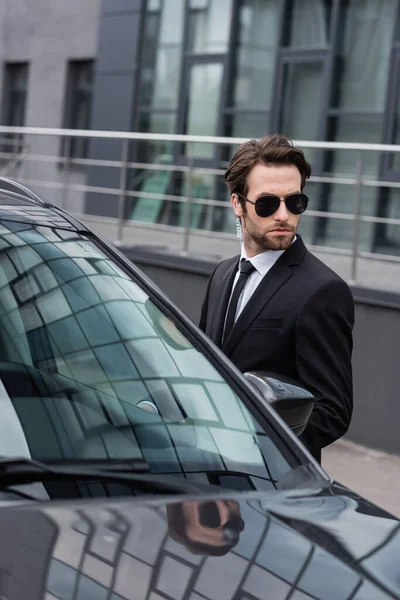 Garde du corps barbu en costume et lunettes de soleil avec oreillette de sécurité près de l'automobile moderne — Photo de stock