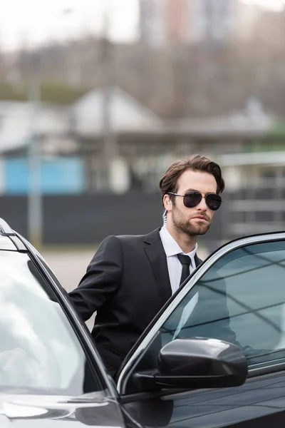 Guardia del corpo barbuta in tuta e occhiali da sole con auricolare di sicurezza seduto in auto moderna — Foto stock