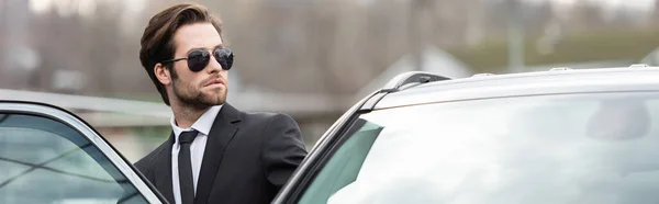 Uomo barbuto in giacca e cravatta e occhiali da sole seduto in auto moderna, banner — Foto stock
