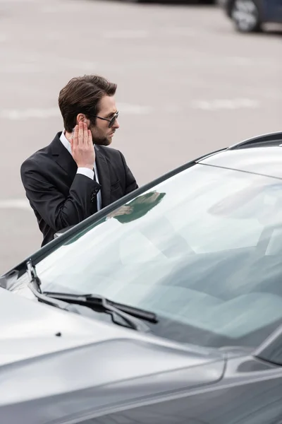 Safeguard in suit and sunglasses adjusting security earpiece near car — Stock Photo