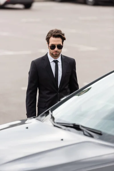 Bodyguard in suit and sunglasses with security earpiece standing near modern auto — Stock Photo