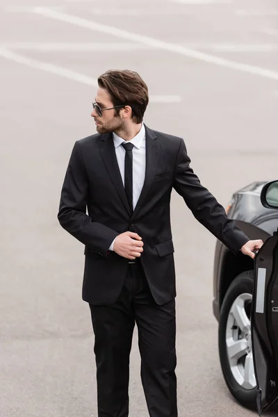 Bearded bodyguard in suit and sunglasses with security earpiece opening door of modern auto — Stock Photo