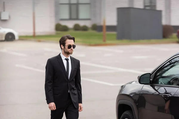 Guardaespaldas barbudo en traje y gafas de sol con auricular de seguridad caminando cerca de auto moderno - foto de stock