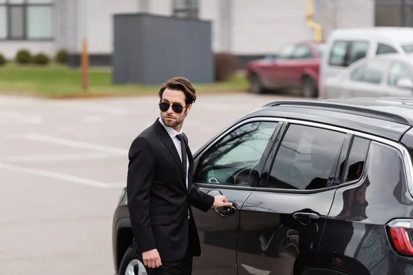 Leibwächter in Anzug und Sonnenbrille mit Sicherheitshörer, der die Tür des modernen Autos öffnet — Stockfoto