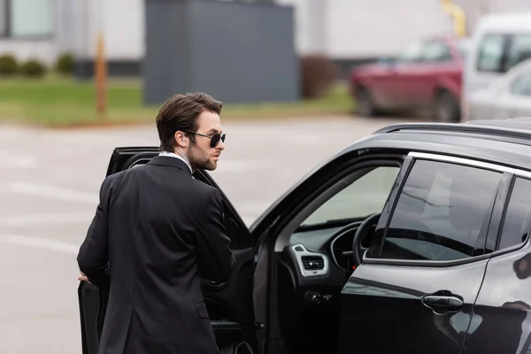 Guardaespaldas barbudo en traje y gafas de sol con auricular de seguridad cerca de la puerta abierta de auto moderno - foto de stock