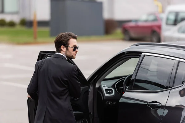 Garde du corps barbu en costume et lunettes de soleil avec oreillette de sécurité près de la porte ouverte de l'automobile moderne — Photo de stock