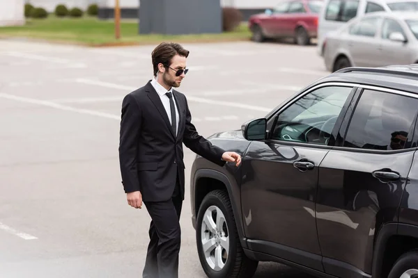 Garde du corps barbu en costume et lunettes de soleil avec écouteur de sécurité marchant près de la voiture moderne — Photo de stock