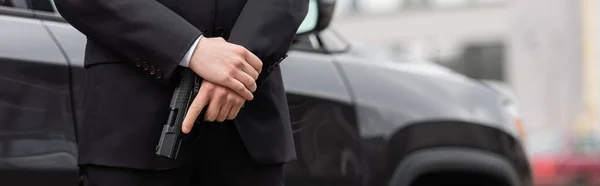 Vista recortada de guardaespaldas en traje con pistola cerca de auto moderno, pancarta - foto de stock