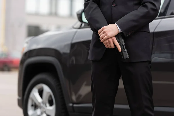 Cropped view of bodyguard in suit holding gun near blurred modern auto — Stock Photo