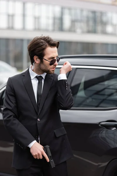 Bodyguard in sunglasses with security earpiece holding gun and talking near modern auto — Stock Photo