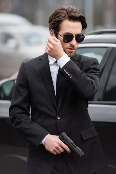 Bearded bodyguard in sunglasses with security earpiece holding gun and talking near modern auto — Stock Photo