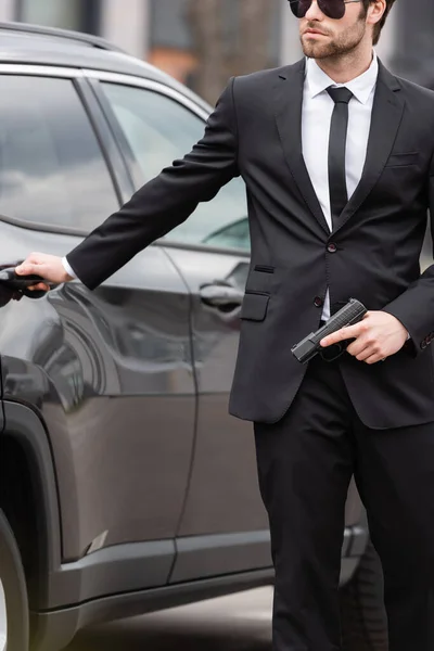 Vista recortada de la salvaguardia en traje y gafas de sol sosteniendo pistola cerca borrosa auto moderno - foto de stock