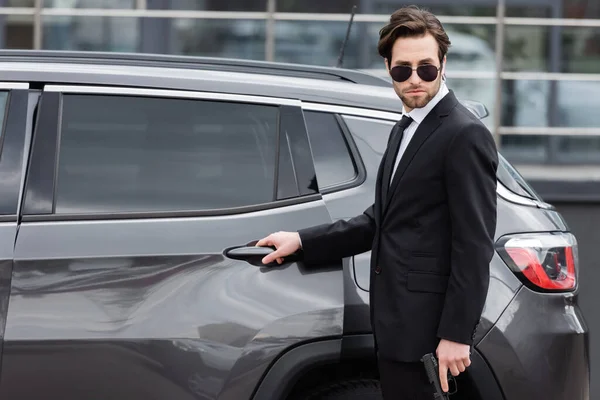 Захист у костюмі та сонцезахисних окулярах та костюмі, що тримає пістолет біля сучасного авто — стокове фото