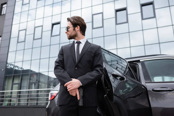 Vue à angle bas du garde du corps barbu en costume tenant pistolet près de la voiture — Photo de stock