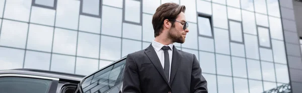 Vista lateral del guardaespaldas barbudo en traje cerca del coche y el edificio moderno, bandera - foto de stock