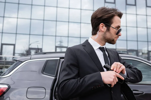 Bearded bodyguard in sunglasses and suit holding gun near modern building — Stock Photo