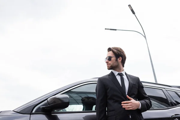 Visão de baixo ângulo de guarda-costas barbudo em óculos de sol e terno de pé perto do carro moderno — Fotografia de Stock