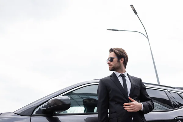 Vista de ángulo bajo del guardaespaldas barbudo en gafas de sol y traje de pie cerca de coche moderno - foto de stock