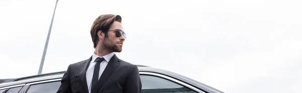 Vista de ángulo bajo del guardaespaldas barbudo en gafas de sol y traje de pie cerca de coche moderno, pancarta - foto de stock