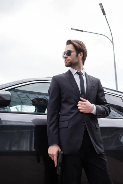 Low angle view of bearded safeguard in sunglasses and suit standing near modern car — Stock Photo