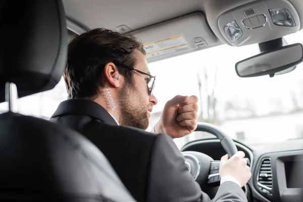 Bärtiger Leibwächter mit Sonnenbrille und Sicherheitshörer, der während der Fahrt durch ein modernes Auto spricht — Stockfoto