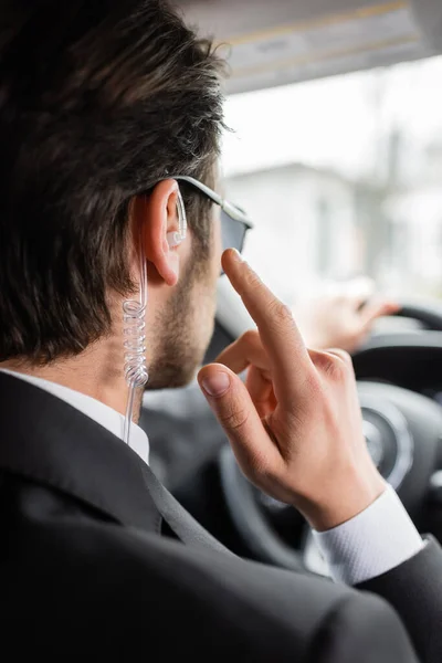 Vista trasera del guardaespaldas en gafas de sol apuntando al auricular de seguridad mientras conduce un automóvil moderno - foto de stock