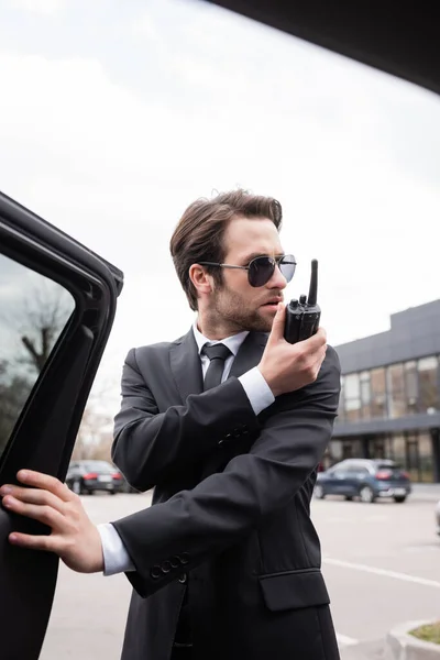 Garde du corps barbu en costume et lunettes de soleil en utilisant talkie walkie près de la voiture — Photo de stock