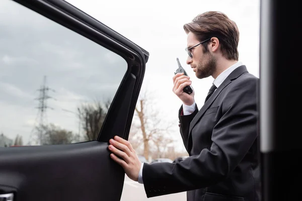 Side view of bearded bodyguard in suit and sunglasses using walkie talkie near modern car — Stock Photo