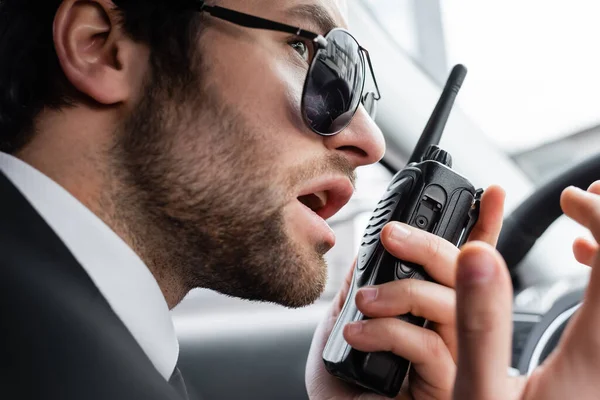 Close up of bearded bodyguard in sunglasses using walkie talkie in car — Stock Photo