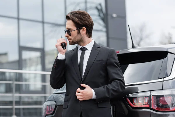 Side view of bodyguard in sunglasses using walkie talkie near modern car — Stock Photo