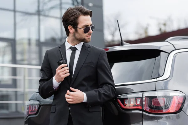 Side view of bodyguard in sunglasses holding walkie talkie near modern car — Stock Photo