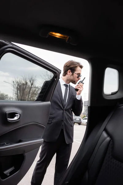 Bodyguard in sunglasses using walkie talkie near modern car — Stock Photo