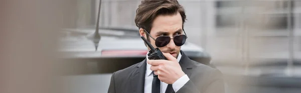 Bodyguard in sunglasses and suit using walkie talkie outside, banner — Stock Photo