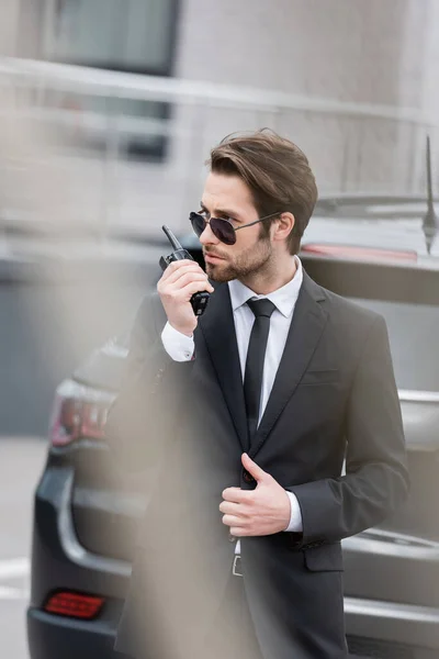 Bodyguard in sunglasses and suit using walkie talkie near blurred modern car — Stock Photo