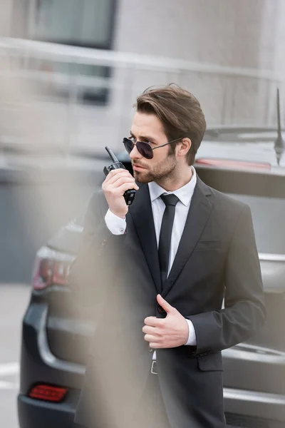 Leibwächter in Sonnenbrille und Anzug mit Walkie Talkie in der Nähe verschwommenes modernes Auto — Stockfoto