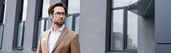 Uomo d'affari barbuto in occhiali e cappotto beige in piedi vicino edificio esterno, banner — Foto stock