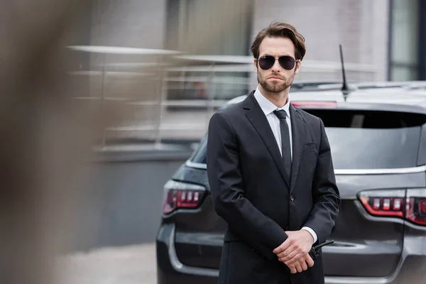 Bodyguard in sunglasses and suit standing with walkie talkie near modern car — Stock Photo