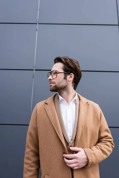 Uomo d'affari barbuto in occhiali e cappotto beige in piedi vicino edificio esterno — Foto stock