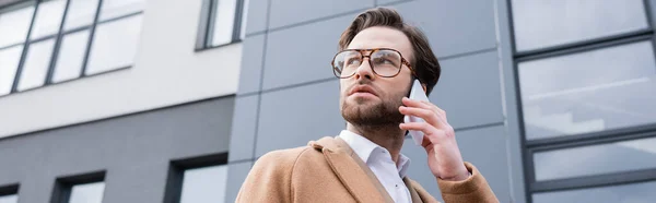 Tiefansicht eines bärtigen Geschäftsmannes mit Brille und beigem Mantel, der in der Nähe von Gebäuden auf dem Handy spricht, Banner — Stockfoto